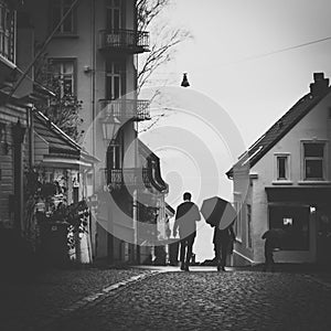 Greyscale shot of people with umbrellas walking along the pathway in an old neighborhood