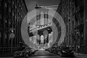 Greyscale of urban buildings and parked cars with Manhattan Bridge in the background