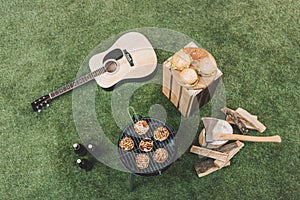 Grill with meat, hamburgers on wooden board, beer bottles and guitar on grass