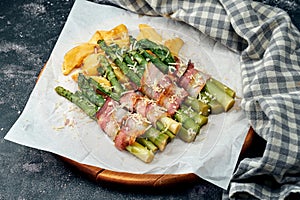 Grilled asparagus with bacon, parmesan cheese and potatoes on white parchment. Dark background