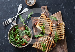 Grilled bacon, mozzarella sandwiches on wooden cutting boards and arugula, cherry tomato salad on dark background, top view.Delici