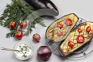 Grilled baked eggplant with tomatoes on metal grill. Dill and tomatoes on table. Sauce in glass bowl