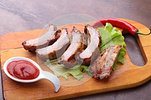 Grilled barbecued ribs with lettuce leaves, hot chili pepper and sauce on wooden cutting board.