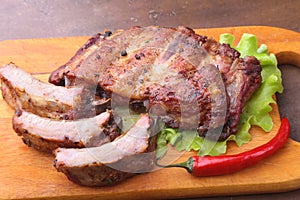 Grilled barbecued ribs with lettuce leaves, hot chili pepper and sauce on wooden cutting board.