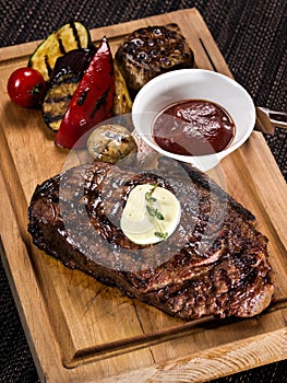 Grilled beef angus steak with melted butter on a wooden cutting board served with grilled vegetables, close up photo on a dark
