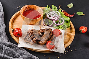 Grilled beef meat and vegetables with fresh salad and bbq sauce on cutting board over black stone background. Hot Meat Dishes