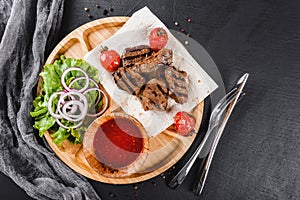 Grilled beef meat and vegetables with fresh salad and bbq sauce on cutting board over black stone background