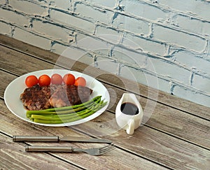 Grilled beef steak with boiled asparagus and cherry tomatoes on a wooden table, cutlery and sauce nearby