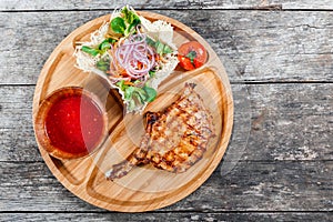 Grilled beef steak on bone, fresh salad, grilled vegetables and tomato sauce on cutting board on wooden background