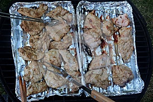 Grilled chicken breast slices placed on alluminium plates on grill, turned by steel forceps and fork during barbecue process.