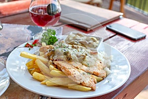 Grilled chicken fillet in white mushroom sauce with french fries on white plate. Glass of red wine in background