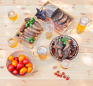 Grilled chicken wings, grilled sausages, beer, bread and bowl with tomatoes on wooden table. Top view