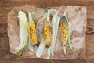 Grilled corn over oily craft paper and rustic wooden background, top view.