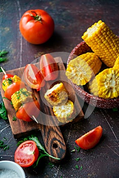 Grilled corn on skewers with tomatoes, herbs and spices on a dark background.