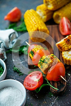 Grilled corn on skewers with tomatoes, herbs and spices on a dark background.