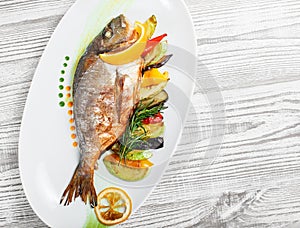 Grilled dorado fish with baked vegetables and rosemary on plate on wooden background close up.