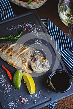 Grilled dorado fish on a plate with vegetables, lemon and a glass of white wine.