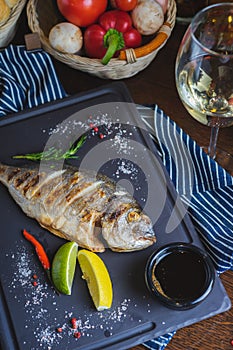 Grilled dorado fish on a plate with vegetables, lemon and a glass of white wine.