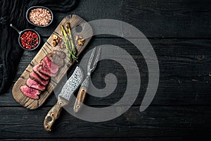 Grilled fillet beef steaks, with onion and asparagus, on wooden serving board, with meat knife and fork, on black wooden table