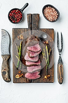 Grilled fillet beef steaks, with onion and asparagus, on wooden serving board, with meat knife and fork, on white stone
