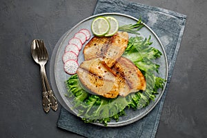 Grilled fish sea bass fillet with green lettuce in a plate on a black stone background