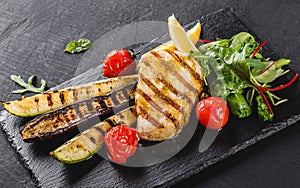 Grilled fish steak garnished with salad of spinach and grilled vegetables on shale background over dark background. Hot fish dish