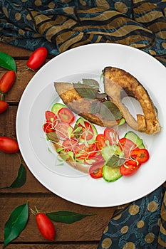 Grilled fish steak with vegetables on plate: tomatoes, microgran, cucumber, tasty and healthy dinner. Wooden rustic background. To