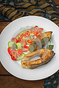 Grilled fish steak with vegetables on plate: tomatoes, microgran, cucumber, tasty and healthy dinner. Wooden rustic background. To