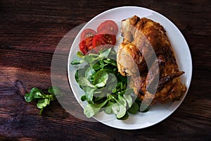 grilled half chicken with tomatoes and corn salad on a white plate on a dark rustic wooden board, top view from above with copy