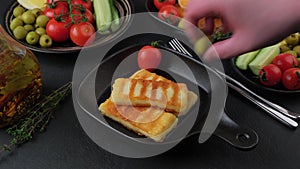 Grilled halloumi cheese in a black pan with olives, tomatoes, cucumbers, thyme and lemon. Man puts olives, tomatoes and lemon.