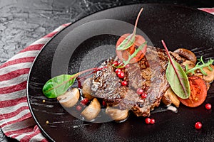 Grilled juicy steak with grilled mushrooms, tomatoes and cranberries on a dark plate and rad napkin on a dark background