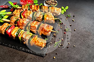 Grilled kebab pieces with vegetables on skewers on a ceramic black board on a dark background with fresh basil leaves