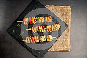 Grilled kebab pieces with vegetables on skewers on a ceramic black board on a dark background with fresh basil leaves