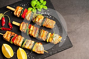 Grilled kebab pieces with vegetables on skewers on a ceramic black board on a dark background with fresh basil leaves