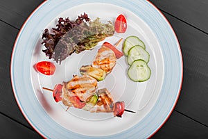 Grilled meat kebabs on skewers with vegetables, cherry tomato, pepper, zucchini in plate on dark wooden background.