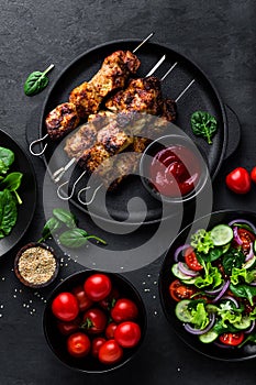 Grilled meat skewers, shish kebab and healthy vegetable salad of fresh tomato, cucumber, onion, spinach, lettuce and sesame on bla