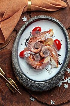Grilled octopus with roasted potatoes, cherry tomatoes in stone bowl on wood board decorated with white flowers.