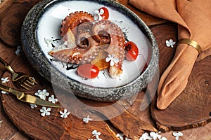 Grilled octopus with roasted potatoes, cherry tomatoes in stone bowl on wood board decorated with white flowers.