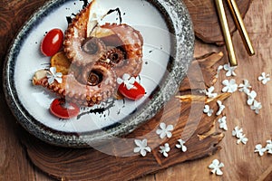 Grilled octopus with roasted potatoes, cherry tomatoes in stone bowl on wood board decorated with white flowers.