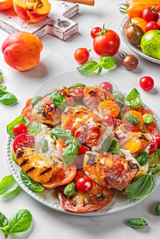 Grilled peaches and prosciutto salad with burrata cheese, tomatoes, cucumber and basil in a plate on white background