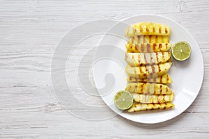 Grilled pineapple slices with lime on white plate and blank notebook over white wooden background, top view. Summer food. From abo