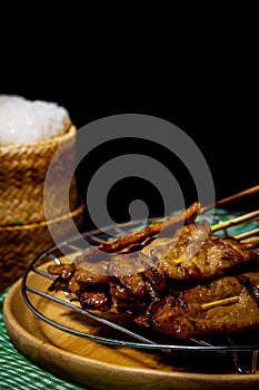 Grilled pork on bamboo skewers placed on a steel grill in a wooden tray with sticky rice in the cart, ready to serve