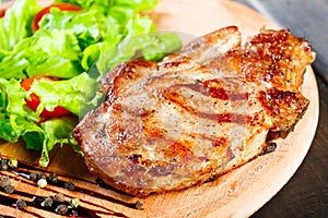 Grilled pork chop steak with fresh vegetable salad, tomatoes and sauce on wooden cutting board. Hot Meat Dishes