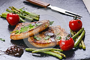 Grilled pork chops, steaks with vegetables, tomatoes, beans and sauce on a black slate. Fresh meat with foam. Dark background. Gri