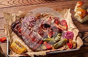 Grilled pork ribs served with grilled corn, salat, bbq sauce, salt pepper and cucumber on parchment paper on a wooden