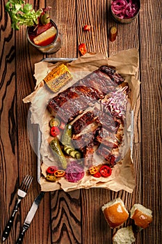 Grilled pork ribs served with grilled corn, salat, bbq sauce, salt pepper and cucumber on parchment paper on a wooden