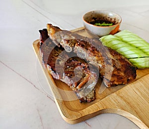 Grilled pork ribs, sliced cucumber and spicy sauce on wooden tray. Close up Barbecue food with fresh vegetables on white marble