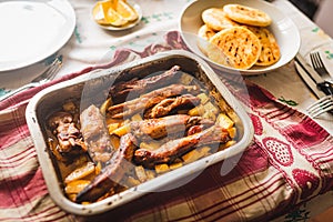 Grilled pork ribs on table inside oven aluminum container with venezuelan arepas and potatoes, table cloth, and warm natural light