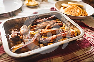 Grilled pork ribs on table inside oven aluminum container with venezuelan arepas and potatoes, table cloth, and warm natural light