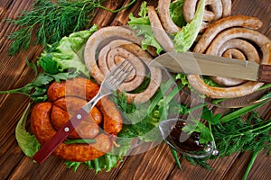 Grilled pork sausages, knife, fork, sauce in a sauce-pan and corn buns on a wooden surface of pine boards. Nearby are lettuce,
